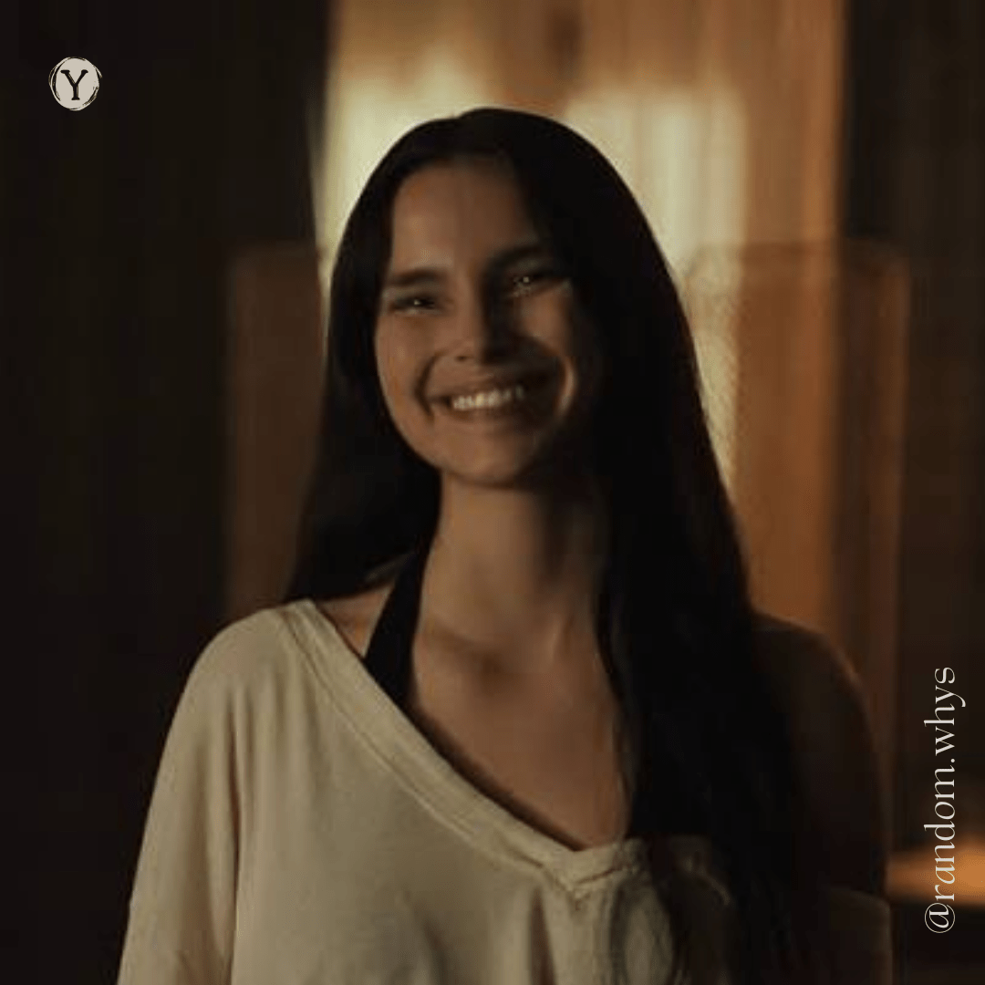 A smiling woman with long dark hair stands indoors in warm lighting, wearing a loose light-colored top with a black strap visible, looking directly at the camera.